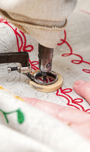Мастер по вышивке работает на вышивальной машине в технике «тамбурный шов». Маленькое пяльце фиксирует ткань, при помощи ручки мастер направляет иглу строго по намеченному контуру